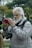 An elderly man with a white beard is capturing a moment with his smartphone outdoors. He is wearing a light grey jacket and has a DSLR camera hanging around his neck. The background features trees and greenery, suggesting a park or nature setting. Another person in the background also holds a smartphone, partially visible.