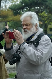 An elderly man with a white beard is capturing a moment with his smartphone outdoors. He is wearing a light grey jacket and has a DSLR camera hanging around his neck. The background features trees and greenery, suggesting a park or nature setting. Another person in the background also holds a smartphone, partially visible.