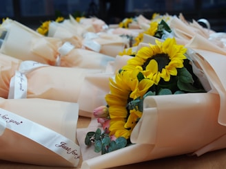A collection of wrapped sunflower bouquets placed on a wooden surface. Each bouquet is wrapped in light peach-colored paper and tied with a white ribbon that reads 'Just for you'. The sunflowers are vibrant with large, yellow petals and prominent dark centers. Smaller pink roses and green foliage are also visible within the bouquets.
