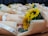 A collection of wrapped sunflower bouquets placed on a wooden surface. Each bouquet is wrapped in light peach-colored paper and tied with a white ribbon that reads 'Just for you'. The sunflowers are vibrant with large, yellow petals and prominent dark centers. Smaller pink roses and green foliage are also visible within the bouquets.