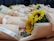 A collection of wrapped sunflower bouquets placed on a wooden surface. Each bouquet is wrapped in light peach-colored paper and tied with a white ribbon that reads 'Just for you'. The sunflowers are vibrant with large, yellow petals and prominent dark centers. Smaller pink roses and green foliage are also visible within the bouquets.