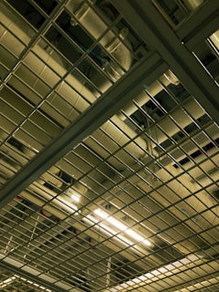 A view of an industrial ceiling with exposed pipes and a grid-like metal framework. Fluorescent lights are visible, casting a muted glow onto the metal surfaces.