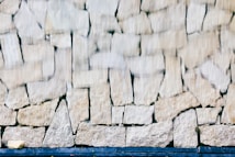A textured stone wall composed of irregular, rectangular stones with a slightly blurred effect, suggesting movement or water flowing over it. The stones are light-colored and have a rough surface, with varying sizes and shapes.