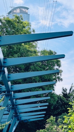 A modern structure with blue steel beams is set against a backdrop of lush green trees and a blue sky. Above, a skyscraper with reflective glass is partially visible, along with several power lines cutting through the sky.