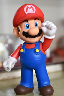 A colorful figurine of a cartoon character wearing a red hat with an M emblem, blue overalls, a red shirt, white gloves, and brown shoes. The character has a prominent black mustache and large blue eyes.