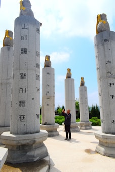 Tall stone pillars are inscribed with Chinese characters and topped with sculptures of animals, featuring a man taking pictures in the center surrounded by greenery and a blue sky.