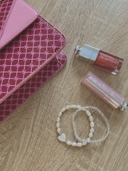 A pink patterned clutch with a geometric design lies on a wooden surface. Nearby are two lip glosses, one with a pink hue and the other with clear packaging. A set of pearl and transparent beaded bracelets, one featuring a heart charm, is also present.