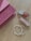 A pink patterned clutch with a geometric design lies on a wooden surface. Nearby are two lip glosses, one with a pink hue and the other with clear packaging. A set of pearl and transparent beaded bracelets, one featuring a heart charm, is also present.
