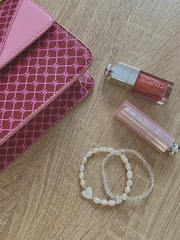 A pink patterned clutch with a geometric design lies on a wooden surface. Nearby are two lip glosses, one with a pink hue and the other with clear packaging. A set of pearl and transparent beaded bracelets, one featuring a heart charm, is also present.