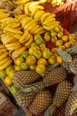 A vibrant assortment of tropical fruits is displayed on a market stand, featuring clusters of bananas, oranges, and pineapples. The bananas are bunched together with a rich yellow hue, while the oranges are a mix of green and orange colors. The pineapples, with their textured skin and leafy tops, are arranged at the bottom of the display.