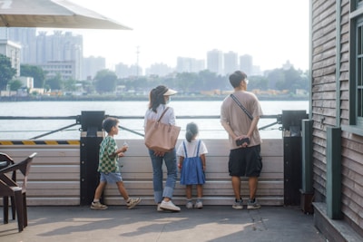A family of four stands on a waterfront, facing the water. The setting appears to be urban with a cityscape in the background, featuring high-rise buildings. The family consists of two adults and two children. The woman is wearing a hat and carrying a light pink bag, the man has his hands behind his back and is carrying a camera. The two children are facing the water, with one of them wearing blue overalls. The scene is set during the day and there is a wooden building on the right side of the image.