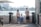 A family of four stands on a waterfront, facing the water. The setting appears to be urban with a cityscape in the background, featuring high-rise buildings. The family consists of two adults and two children. The woman is wearing a hat and carrying a light pink bag, the man has his hands behind his back and is carrying a camera. The two children are facing the water, with one of them wearing blue overalls. The scene is set during the day and there is a wooden building on the right side of the image.