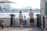 A family of four stands on a waterfront, facing the water. The setting appears to be urban with a cityscape in the background, featuring high-rise buildings. The family consists of two adults and two children. The woman is wearing a hat and carrying a light pink bag, the man has his hands behind his back and is carrying a camera. The two children are facing the water, with one of them wearing blue overalls. The scene is set during the day and there is a wooden building on the right side of the image.