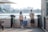 A family of four stands on a waterfront, facing the water. The setting appears to be urban with a cityscape in the background, featuring high-rise buildings. The family consists of two adults and two children. The woman is wearing a hat and carrying a light pink bag, the man has his hands behind his back and is carrying a camera. The two children are facing the water, with one of them wearing blue overalls. The scene is set during the day and there is a wooden building on the right side of the image.