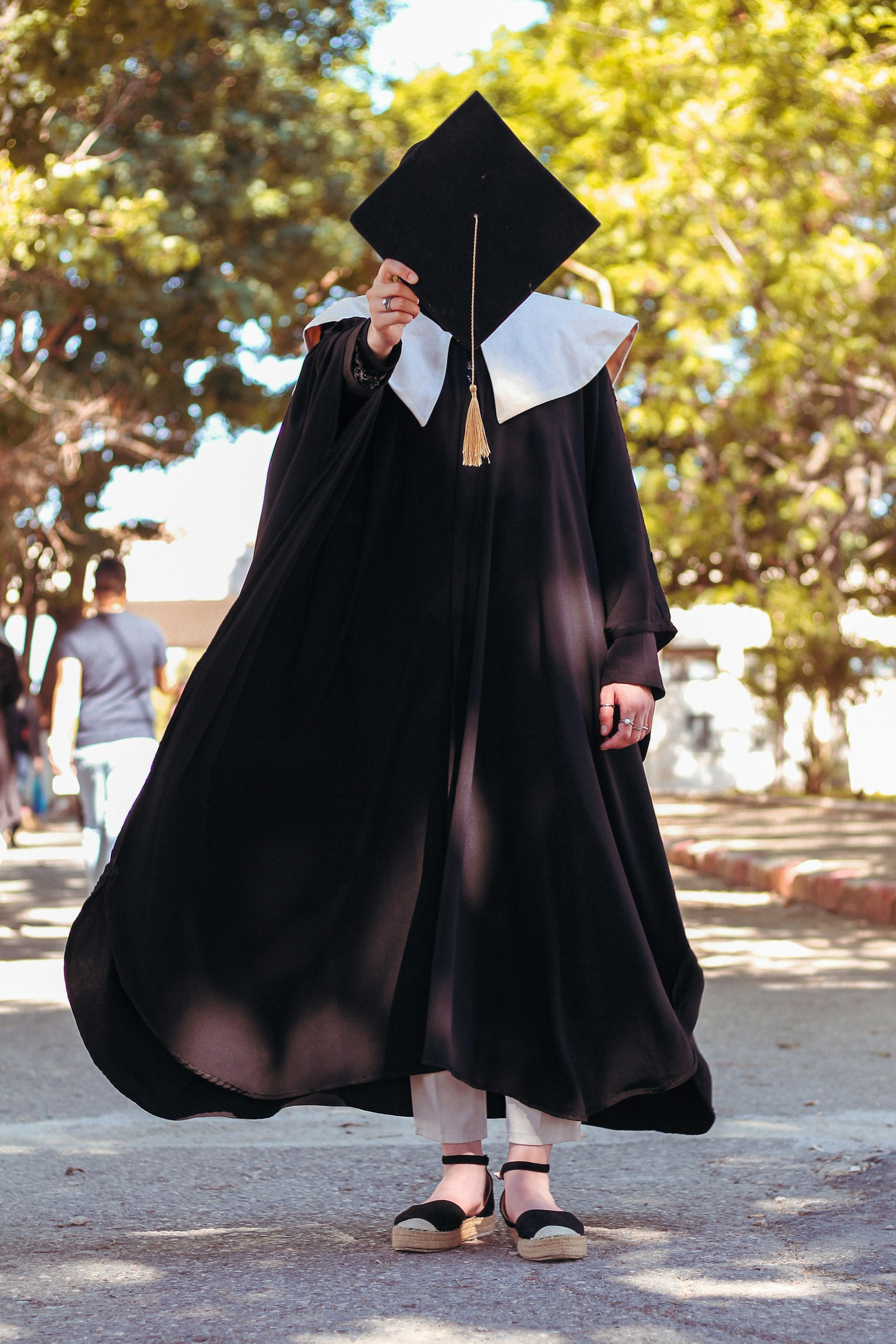 A person in a black graduation gown holds a mortarboard cap in front of their face while standing outdoors. The scene is set in a leafy area with trees and a sunny ambiance. The gown flows down gracefully, and the mortarboard has a gold tassel.
