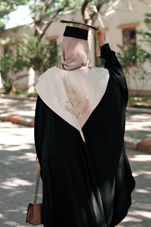 A person wearing a graduation gown and cap stands outside in a leafy environment. The cap is held in one hand, and a cream-colored stole is draped over the shoulders. The sunlight filters through the trees, casting dappled shadows on the path.