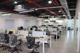 A modern open-plan office space features several desks with office chairs and computer monitors. The room has a sleek design with glass partitions and plants hanging from the ceiling. Flags and organized workspaces are visible, with whiteboards and notes on the walls.
