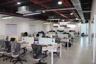 A modern open-plan office space features several desks with office chairs and computer monitors. The room has a sleek design with glass partitions and plants hanging from the ceiling. Flags and organized workspaces are visible, with whiteboards and notes on the walls.