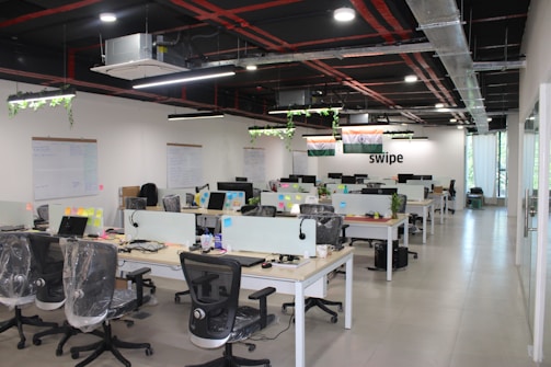 A modern open-plan office space features several desks with office chairs and computer monitors. The room has a sleek design with glass partitions and plants hanging from the ceiling. Flags and organized workspaces are visible, with whiteboards and notes on the walls.
