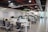 A modern open-plan office space features several desks with office chairs and computer monitors. The room has a sleek design with glass partitions and plants hanging from the ceiling. Flags and organized workspaces are visible, with whiteboards and notes on the walls.