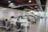 A modern open-plan office space features several desks with office chairs and computer monitors. The room has a sleek design with glass partitions and plants hanging from the ceiling. Flags and organized workspaces are visible, with whiteboards and notes on the walls.