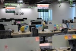 A modern office space with rows of desks, each equipped with computer monitors and office chairs. There are Indian flags hanging from the ceiling. The desks have various items on them, including sticky notes, water bottles, and some personal items. Plants are hanging from the ceiling, adding a touch of greenery. The word 'swipe' is displayed on the wall.
