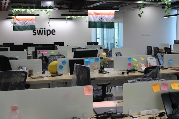 A modern office space with rows of desks, each equipped with computer monitors and office chairs. There are Indian flags hanging from the ceiling. The desks have various items on them, including sticky notes, water bottles, and some personal items. Plants are hanging from the ceiling, adding a touch of greenery. The word 'swipe' is displayed on the wall.