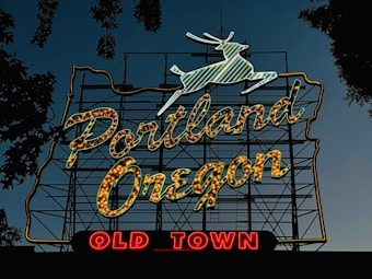 A large neon sign featuring a leaping white stag situated above the words 'Portland Oregon' in glowing yellow-orange. Below, 'Old Town' is displayed in bright red neon. The sign is set against a twilight sky, with silhouettes of tree branches framing the edges.