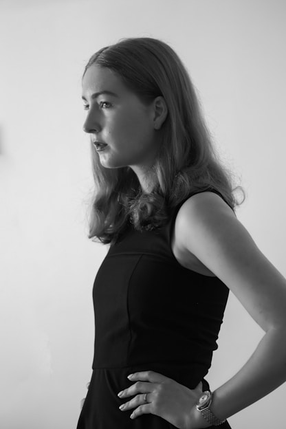 A black and white portrait of a person with long hair standing in profile, wearing a sleeveless dark dress. The background is plain and minimalistic, enhancing the focus on the subject.