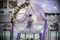 A beautifully decorated outdoor setting features a central three-tiered cake adorned with purple accents. Surrounding the cake is a floral arch with white and purple flowers, enhanced by a delicate purple drapery. The background reveals a serene poolside ambience with sparkling lights adding to the elegance.