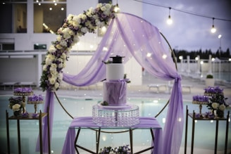 A beautifully decorated outdoor setting features a central three-tiered cake adorned with purple accents. Surrounding the cake is a floral arch with white and purple flowers, enhanced by a delicate purple drapery. The background reveals a serene poolside ambience with sparkling lights adding to the elegance.