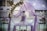 A beautifully decorated outdoor setting features a central three-tiered cake adorned with purple accents. Surrounding the cake is a floral arch with white and purple flowers, enhanced by a delicate purple drapery. The background reveals a serene poolside ambience with sparkling lights adding to the elegance.