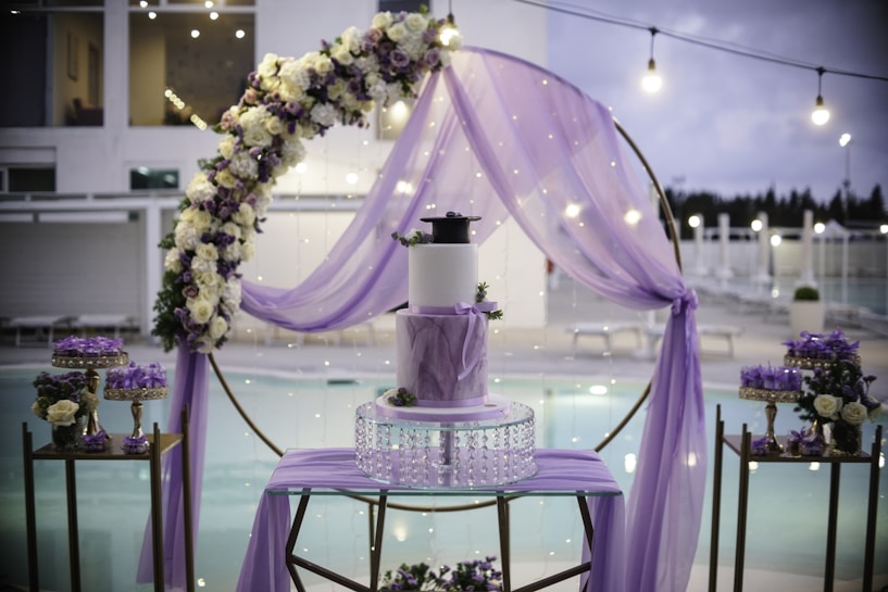 A beautifully decorated outdoor setting features a central three-tiered cake adorned with purple accents. Surrounding the cake is a floral arch with white and purple flowers, enhanced by a delicate purple drapery. The background reveals a serene poolside ambience with sparkling lights adding to the elegance.