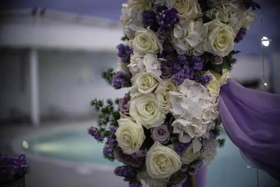 A decorative floral arrangement featuring an assortment of white roses and purple flowers, possibly lavender. This elegant bouquet is accompanied by lush green foliage and draped with light purple fabric. The background shows a blurred view of an indoor or outdoor setting with a pool, creating a serene ambiance.