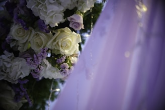 A close-up of an elegantly arranged floral decoration featuring white and pale purple roses intertwined with green leaves. The delicate flowers are adorned with small fairy lights, and the background fades into soft hues of lavender.