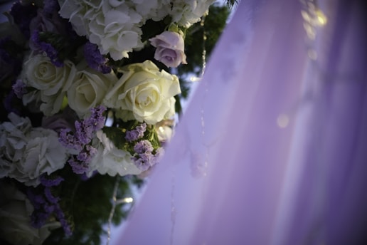 A close-up of an elegantly arranged floral decoration featuring white and pale purple roses intertwined with green leaves. The delicate flowers are adorned with small fairy lights, and the background fades into soft hues of lavender.