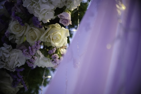 A close-up of an elegantly arranged floral decoration featuring white and pale purple roses intertwined with green leaves. The delicate flowers are adorned with small fairy lights, and the background fades into soft hues of lavender.