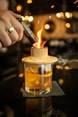 A handcrafted cocktail is being prepared with a torch igniting a wooden lid over the glass. Inside the glass, there is an orange slice and a cherry, suggesting a smoky flavor infusion. The setting is elegantly lit, with warm lights creating a cozy and sophisticated atmosphere.