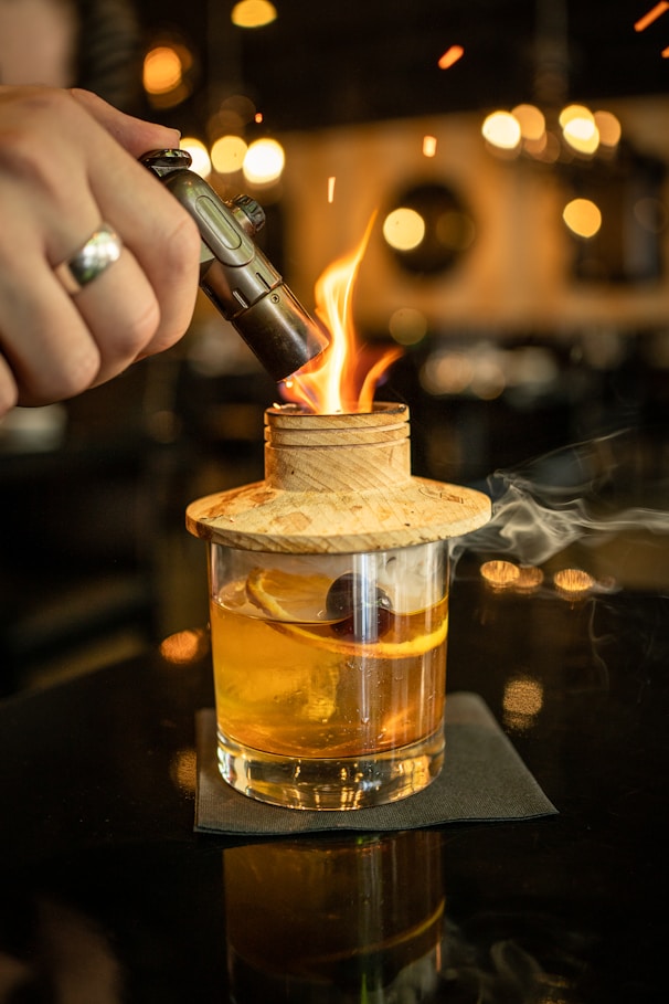 A handcrafted cocktail is being prepared with a torch igniting a wooden lid over the glass. Inside the glass, there is an orange slice and a cherry, suggesting a smoky flavor infusion. The setting is elegantly lit, with warm lights creating a cozy and sophisticated atmosphere.