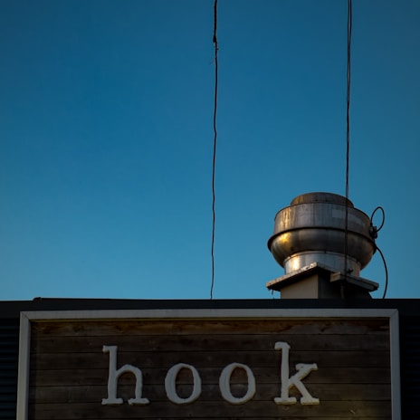 A wooden sign with the word 'hook' is prominently displayed against a clear blue sky. Above the sign, an industrial metal vent with wires is visible, giving a rustic and somewhat urban appearance.