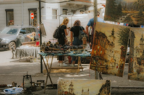 A street market scene with various paintings on display in the foreground. The artwork features urban landscapes with historical buildings. Several people are browsing the market, examining items or paintings on tables. The atmosphere appears casual and relaxed, with soft and warm lighting that suggests a sunny day.