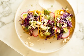 Three tacos are arranged on a light-colored plate. They are topped with vibrant ingredients such as shredded purple cabbage, radish slices, avocado chunks, crumbled cheese, and garnished with fresh herbs. The plate is set on a marble surface.
