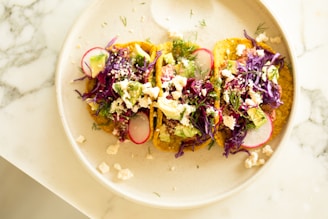 Three tacos are arranged on a light-colored plate. They are topped with vibrant ingredients such as shredded purple cabbage, radish slices, avocado chunks, crumbled cheese, and garnished with fresh herbs. The plate is set on a marble surface.