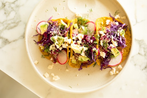 Three tacos are arranged on a light-colored plate. They are topped with vibrant ingredients such as shredded purple cabbage, radish slices, avocado chunks, crumbled cheese, and garnished with fresh herbs. The plate is set on a marble surface.