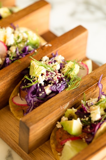 Tacos filled with vibrant ingredients like purple cabbage, avocado, radish slices, feta cheese, sesame seeds, and fresh dill, served in wooden taco holders.