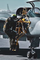A pilot wearing a helmet and a flight suit ascends a yellow ladder to enter the cockpit of a military jet. The aircraft is painted in shades of gray, and the open cockpit canopy reveals its intricate controls.