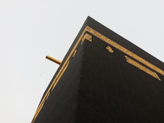 A close-up view of the Kaaba, showcasing its black fabric covering with intricate golden embroidery and calligraphy. The angle highlights the corner of the structure against a pale sky, with a small distant bird also visible.