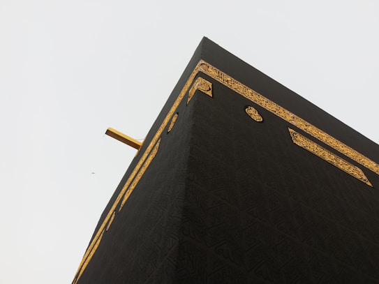 A close-up view of the Kaaba, showcasing its black fabric covering with intricate golden embroidery and calligraphy. The angle highlights the corner of the structure against a pale sky, with a small distant bird also visible.