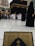 The image depicts a busy scene near the Kaaba in Mecca, with numerous people dressed in traditional pilgrimage attire, such as white robes for men and black coverings for women. In the foreground, a book with ornate gold and black patterns is partially visible, possibly a Quran. Surrounding the Kaaba are large buildings with modern architectural design.
