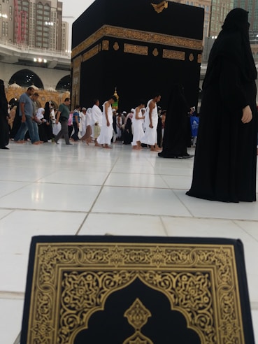 The image depicts a busy scene near the Kaaba in Mecca, with numerous people dressed in traditional pilgrimage attire, such as white robes for men and black coverings for women. In the foreground, a book with ornate gold and black patterns is partially visible, possibly a Quran. Surrounding the Kaaba are large buildings with modern architectural design.